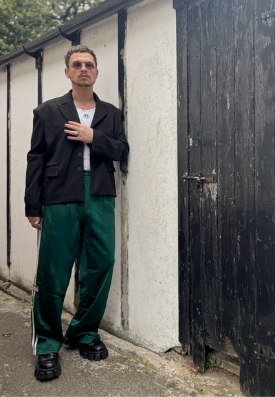 Blazer negro oversized, camiseta blanca, pantalones de satén verdes con franjas laterales, zapatos de plataforma negros y gruesos, de pie al lado de una puerta de madera.