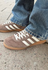 Καφέ σουέντ αθλητικά παπούτσια Adidas με λευκές ρίγες και κορδόνια, φορεμένα με μπλε τζιν, σε υφή οδοστρώμα τος.