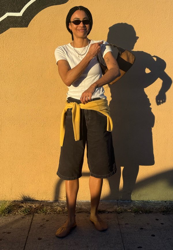 Person med solbriller, hvit t-skjorte, svarte shorts, gul genser bundet rundt livet, holder en brun og svart veske over skulderen foran en gul vegg.
