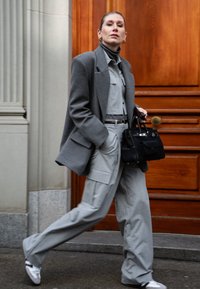 Grå ullblazer över en ljusgrå jumpsuit med fickor, tillsammans med en svart handväska och vita sneakers. Släta texturer, skräddarsydd passform.