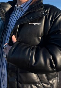 Personne portant une veste en cuir noire avec le logo "lowlights", une chemise à rayures avec col en-dessous, et plusieurs bagues en argent à leurs doigts.