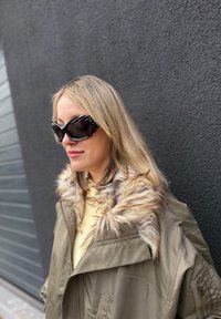 Femme aux cheveux blonds portant de grandes lunettes de soleil noires et un manteau beige avec un col en fourrure, debout contre un mur sombre texturé.