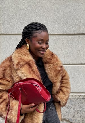 Rød læderhåndtaske med rund form og sidelukning, holdt op mod en struktureret beige pelsfrakke. Guldøreringe komplementerer looket.