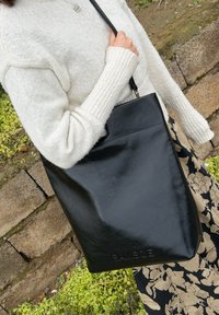 Personne portant un pull en maille blanc et une jupe fleurie, tenant un sac fourre-tout en cuir noir en plein air près d'un mur en pierre et de verdure.