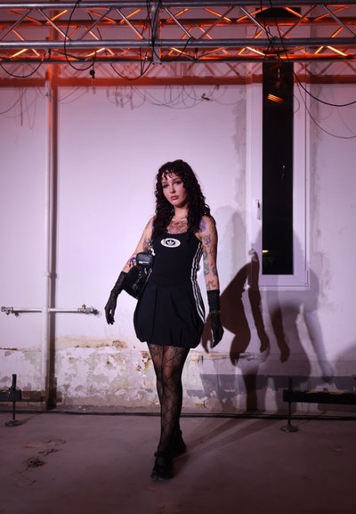 Vestido negro sin mangas con franjas blancas en los lados, guantes de encaje texturizados, medias de red con patrón y bolso pequeño; detalle de fondo mínimo.