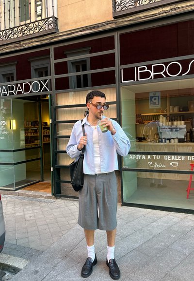 Camisa de rayas azul claro de manga corta; pantalones cortos grises con pliegues; calcetines blancos; mocasines negros; sosteniendo una bebida verde, junto a la entrada de un café.