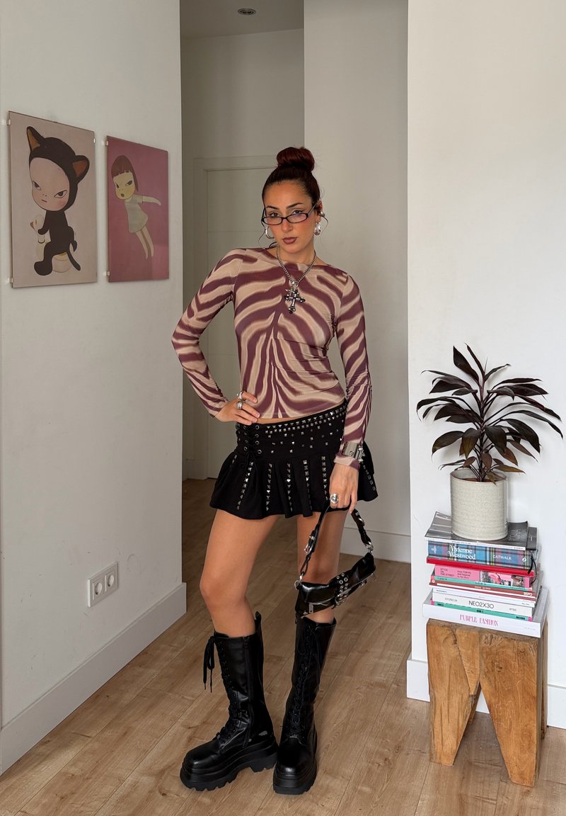 Femme portant un haut à imprimé zèbre, une jupe noire cloutée, de grandes bottes noires, tenant un sac à main noir, debout dans une pièce avec de l'art mural et une plante posée sur des livres.