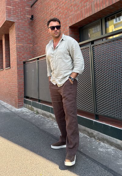 Camisa de lino en beige claro, corte relajado; pantalones de lino marrones; zapatillas grises; gafas de sol grandes; fondo de pared de ladrillo texturizado.