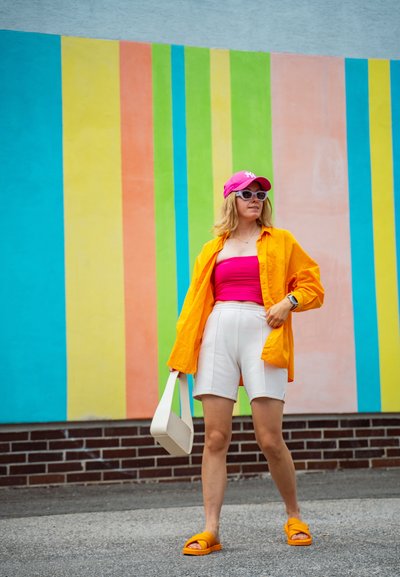 Camisa naranja brillante sobre una camiseta rosa, combinada con pantalones cortos blancos y sandalias naranjas. Los accesorios incluyen una gorra rosa y un bolso blanco. El fondo presenta rayas verticales coloridas.