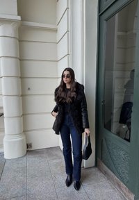 Črni usnjeni plašč z ovratnikom iz krzna, črna majica, temno modre kavbojke in črni gležnjarji zašiljenih konic. Drži majhno teksturirano torbico.