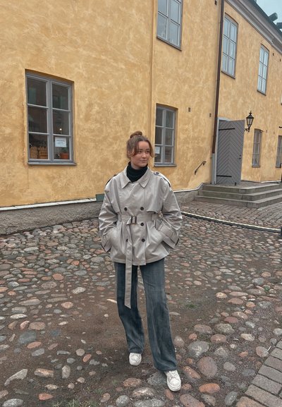 Gabardina beis de doble botonadura con cintura ceñida por un cinturón, combinada con pantalones de pierna ancha gris oscuro y zapatillas blancas, de pie sobre un pavimento de adoquines.