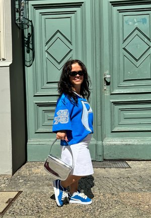 Jersey azul oversize con el número "1973" en la manga, falda blanca, bolso plateado y zapatillas azules con cordones blancos, todo enmarcado por puertas verdes.