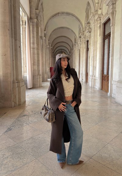Joven mujer con vaqueros anchos, top corto beige, abrigo largo oscuro y gorra gris, de pie en un pasillo arqueado de piedra sosteniendo un bolso estampado.