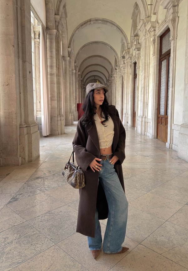 Jonge vrouw in wijde spijkerbroek, beige cropped top, donkeren lange jas en grijze pet staat in hal met stenen bogen terwijl ze een handtasje met patroon vasthoudt.