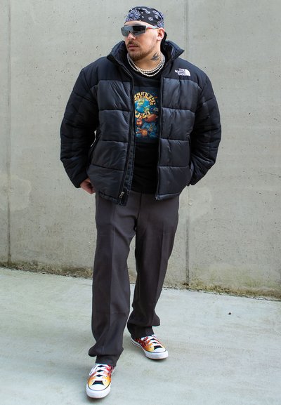 Chaqueta de plumas negra, camiseta negra con gráfico, pantalones grises, zapatillas multicolores. Los accesorios incluyen gafas de sol y una bandana estampada.