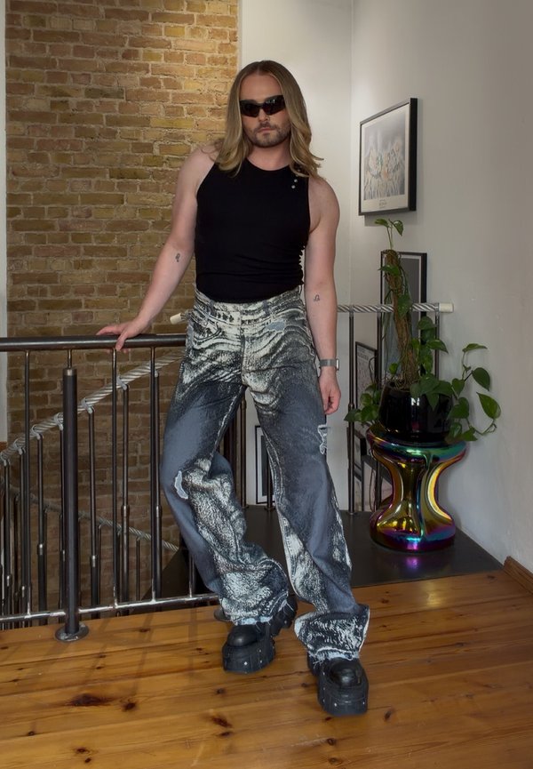 Personne aux longs cheveux portant des lunettes de soleil, un haut noir sans manches, un pantalon large à motifs et des chaussures à plateforme, appuyée sur une rambarde à l'intérieur.