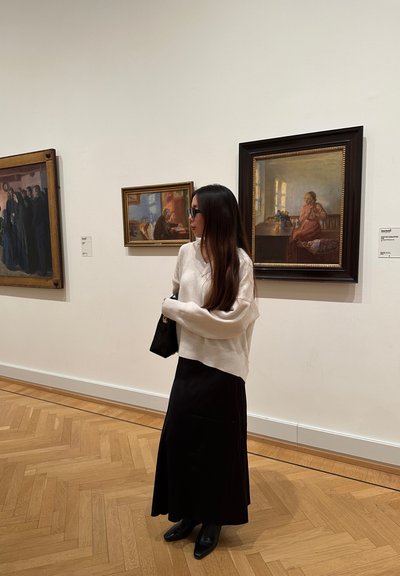 Suéter blanco de gran tamaño con tejido texturizado, falda maxi negra y botas negras de tacón. Rodeada de pinturas enmarcadas en una galería.