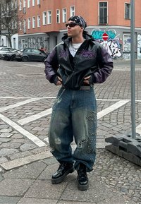 Veste bomber en cuir noir avec des accents violets, pantalon en denim usé avec des embellissements, bottes de combat noires et un bandana.