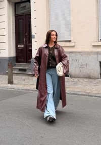 Lange kastanjebruine leren jas, zwarte cardigan, lichtblauwe wijde jeans en een witte tas. Wandelt op een straat met een gestructureerd kinderkopjesoppervlak.