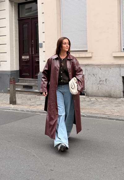 Largo abrigo de cuero burdeos, cardigans negros, pantalones vaqueros de pierna ancha de color azul claro y bolso blanco. Caminando por una calle con una superficie de adoquines texturizados.