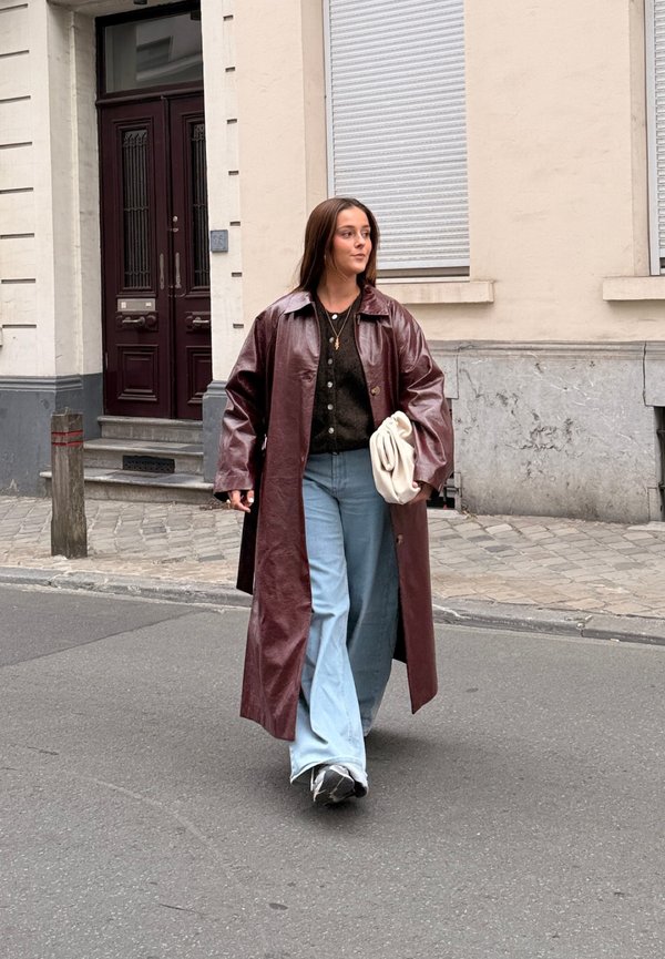 Long manteau en cuir bordeaux, cardigan noir, jeans larges bleu clair et sac blanc. Marchant sur une rue avec une surface en pavés texturés.