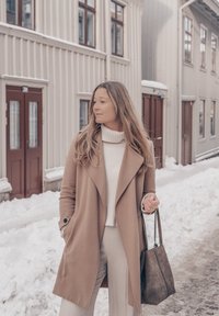 Femme en manteau beige et pull blanc tenant un sac fourre-tout gris, debout dans une rue enneigée bordée de bâtiments en bois beige.