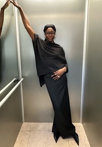 Čierny oversized top a dlhá sukňa z jemnej látky, doplnené o špicaté čierne opätky. Doplnky obsahujú veľké náušnice a šatku na hlave.