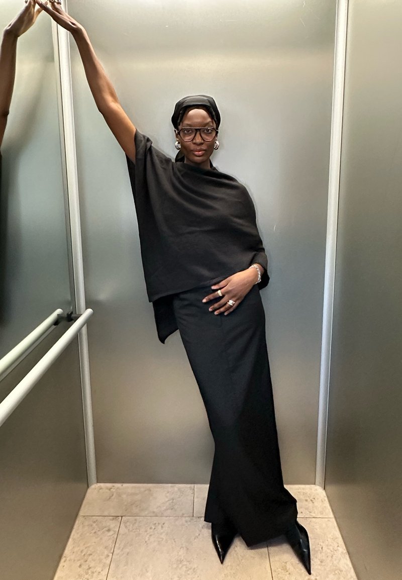 Black oversized top and long skirt in a soft fabric, paired with pointed black heels. Accessorized with large earrings and a headscarf.
