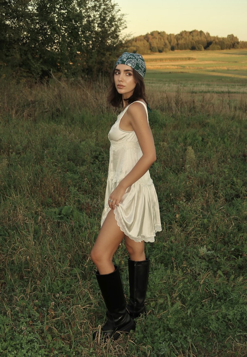 Vestido de seda com acabamento em renda, usado com um lenço verde estampado e botas pretas até ao joelho, situado num campo gramado sob uma luz suave.