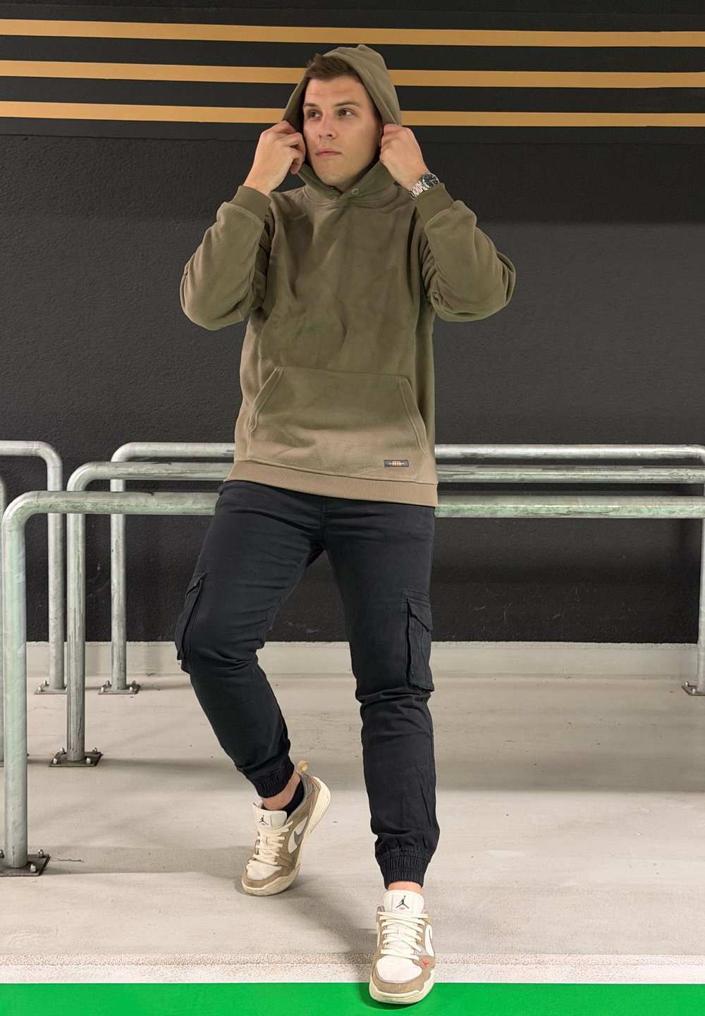 Hanorac cu glugă verde măsliniu, cu buzunar frontal, combinat cu pantaloni negri cargo și adidași bej. Fundalul prezintă pereți gri și balustrade metalice.