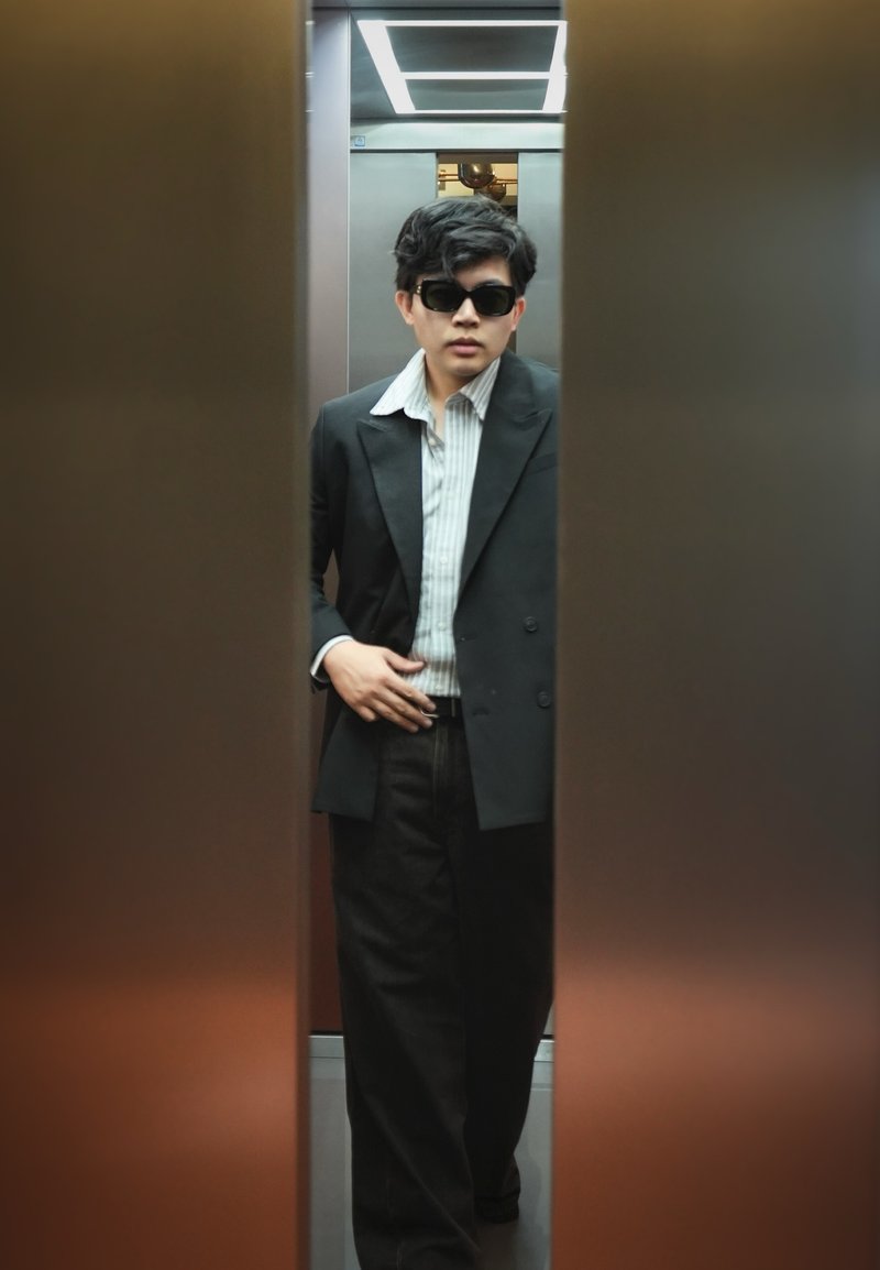 Jeune homme portant des lunettes de soleil, un blazer noir et une chemise rayée debout à l'intérieur d'une porte d'ascenseur partiellement ouverte.