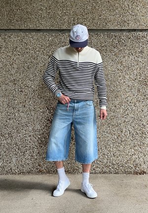 Camisa de rayas de manga larga en crema y azul marino, pantalones cortos de mezclilla anchos y zapatillas blancas. Los accesorios incluyen una gorra y un reloj.