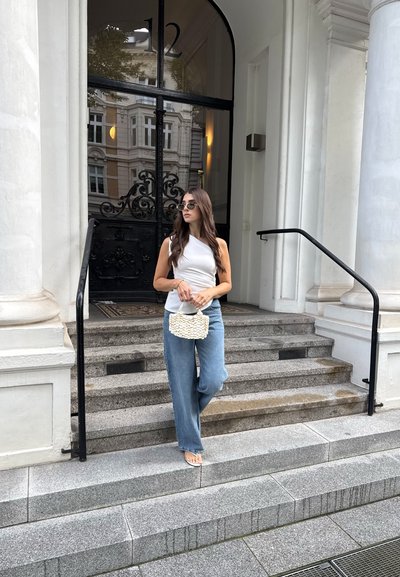 Mujer con top blanco y jeans azules sostiene un bolso blanco, de pie en escalones de piedra frente a un edificio con una puerta negra arqueada y columnas.