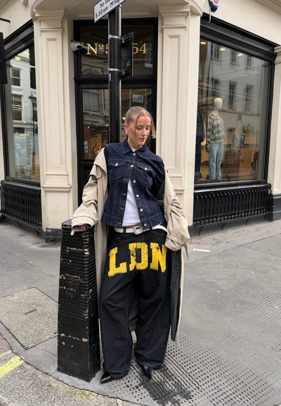 Joven apoyada en un poste negro, vistiendo pantalones amplios con el logo amarillo "LDN", chaqueta vaquera, abrigo beige y zapatos negros puntiagudos en la acera de la ciudad.