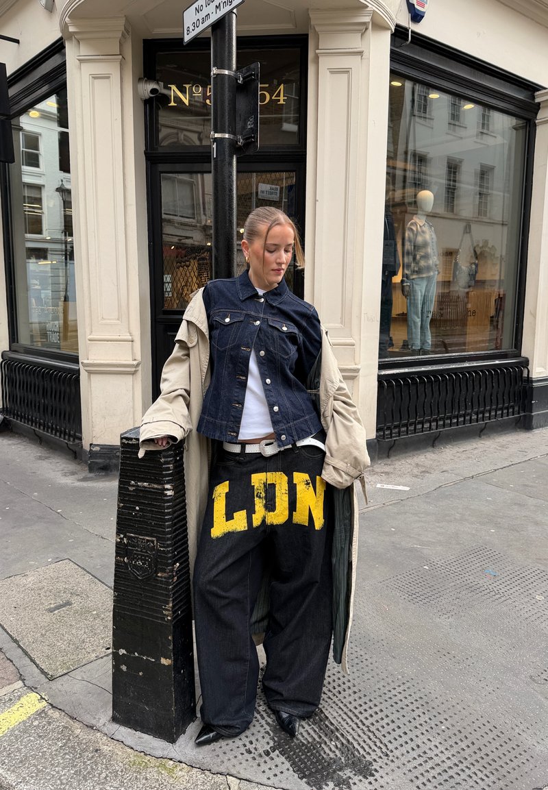Jeune femme appuyée sur un poteau noir, portant un pantalon surdimensionné avec l'inscription jaune « LDN », une veste en denim, un manteau beige et des chaussures noires à bouts pointus sur un trottoir en ville.