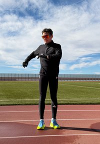 Homme en tenue de sport noire ajustant ses gants sur une piste de course rouge sous un ciel partiellement nuageux.