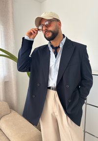 Blazer azul-marinho de dois botões sobre uma camisa riscada azul-claro, combinado com calças creme e um chapéu bege. Óculos de sol castanhos e um cachecol escuro complementam o look.