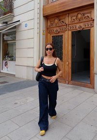 Ženska v črnem topu brez rokavov in modrih visokih jeans hlačah hodi po pločniku blizu lesenih vrat z dekorativnimi dodatki. Rumene čevlje.