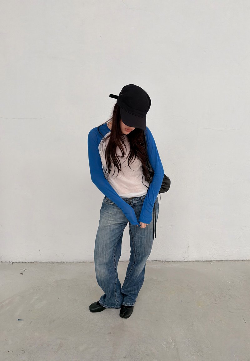 Άτομο με μαύρο καπέλο, μπλε και λευκό μακρυμάνικο πουκάμισο, άνετο μπλε τζιν, μαύρα παπούτσια, στέκεται μπροστά σε έναν απλό λευκό τοίχο.