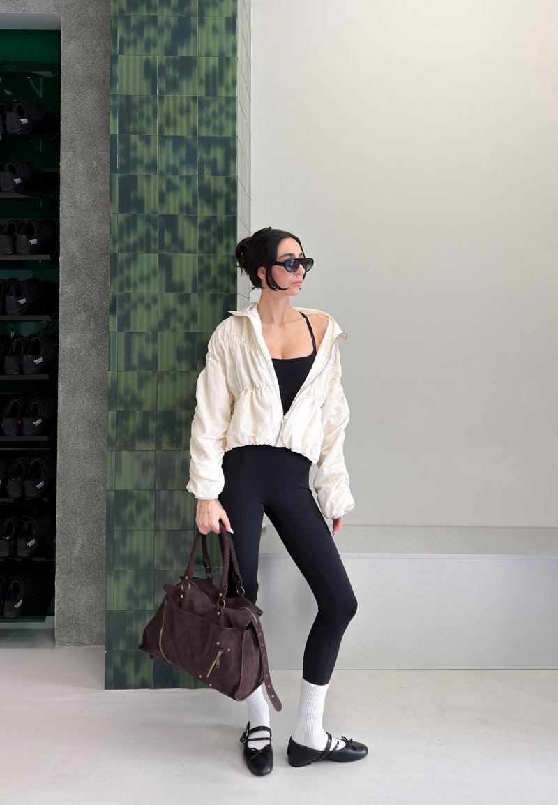 Femme en leggings noirs, veste blanche et lunettes de soleil, tenant un grand sac marron, debout devant un mur composé de carreaux verts et d'une surface unie.