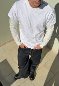 Νέος άνδρας που φοράει στρωμένα λευκά μακρυμάνικα και μπλούζα, μαύρα τζιν και μαύρες μπότες, στέκεται σε πλακόστρωτο πάτωμα στο φως του ήλιου.