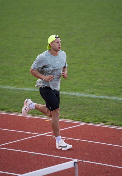 Corredor en una pista, vistiendo una camiseta gris claro con un estampado, pantalones cortos negros, una gorra amarillo neón y zapatillas blancas. Césped de fondo.