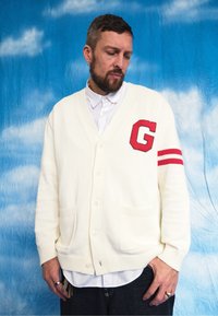 Off-white cardigan featuring a G patch, red stripes on the sleeves, a button-down front, and two front pockets, styled over a white collared shirt.
