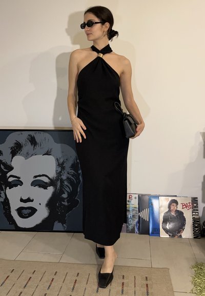 Mujer con un vestido halter negro, gafas de sol negras y tacones, sosteniendo un bolso negro, de pie cerca de una obra de arte de Marilyn Monroe y discos de vinilo.