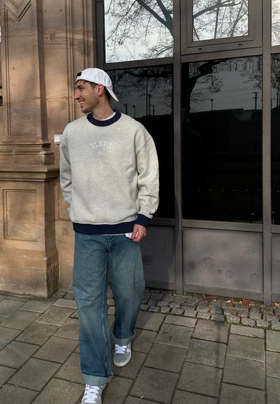 Sudadera gris con acentos en azul marino y texto "BLKVIS." Llevada con unos vaqueros azules holgados y zapatillas blancas, de pie contra un fondo de cristal.