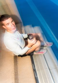 Homme en tenue de sport assis sur les marches au bord de la piscine tenant une bouteille d'eau, avec un effet de flou de mouvement et de l'eau bleue à proximité.