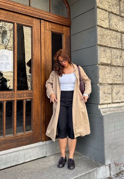 Mujer con gabardina beige, camiseta blanca, pantalones negros recortados y gafas, de pie junto a una puerta de madera con fondo de pared de piedra.