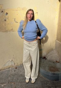 Svetlo modra puhasta majica z napihnjenimi rokavi, združena s kremastimi širokimi hlačami in bež supergami. V ozadju je teksturirana stena.