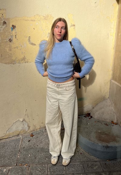 Suéter azul claro y esponjoso con mangas abullonadas, combinado con pantalones anchos crema y zapatillas beige. El fondo presenta una pared texturizada.