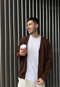 Cardigã castanho de malha sobreposto a uma camisa branca, com calças riscas verticais. Segurando uma chávena vermelha, encostado a um fundo de ripas de madeira.